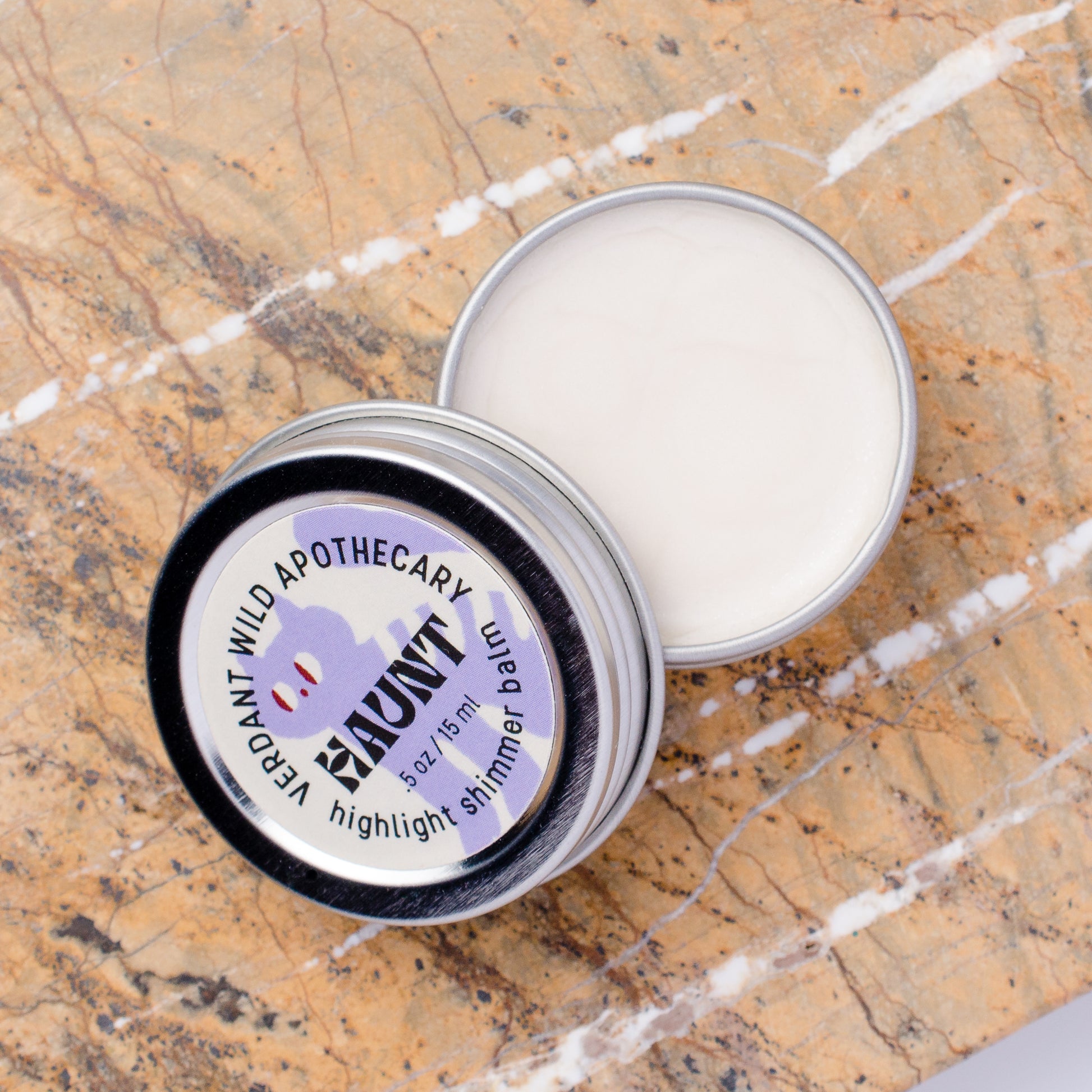 An open silver metal tin of highlighter shimmer balm lies on a brown and white marble surface, with the lid placed beside it. 