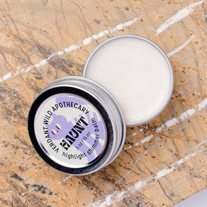 An open silver metal tin of highlighter shimmer balm lies on a brown and white marble surface, with the lid placed beside it. 
