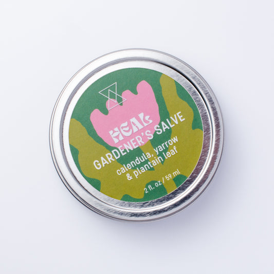 A round silver metal tin of HEAL Gardener's Salve, featuring a colorful label with a pink flower and white text, photographed on a clean white background.