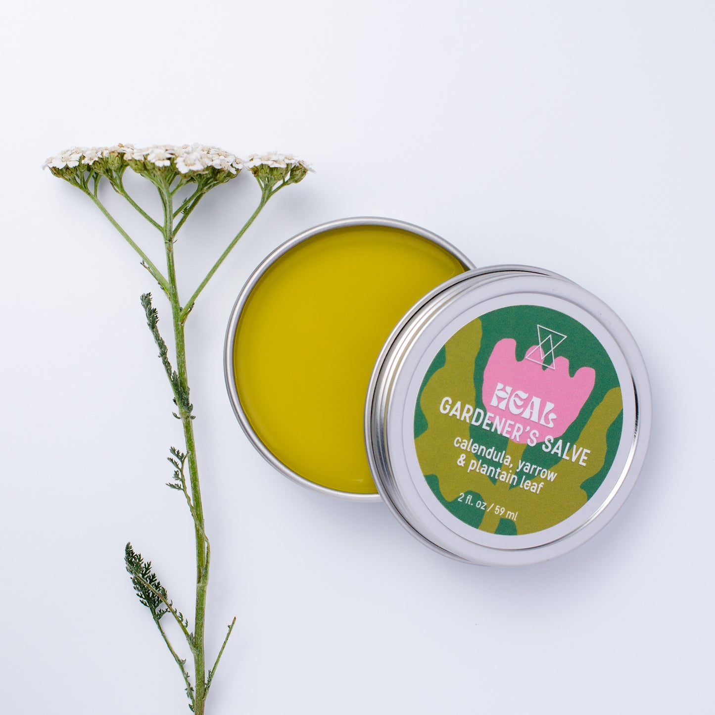 An open round metal tin of HEAL Gardener's Salve showing bright yellow-green balm inside, with the lid leaning against it. The label reads 'HEAL GARDENER'S SALVE' and a sprig of yarrow decorates the image.