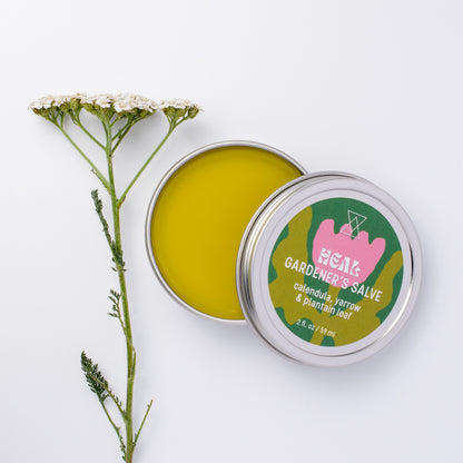 An open round metal tin of HEAL GARDENER'S SALVE showing the yellow-green balm inside, with the lid placed beside it. The lid label is green and pink with white text reading 'HEAL GARDENER'S SALVE' and a sprig of yarrow off to side.