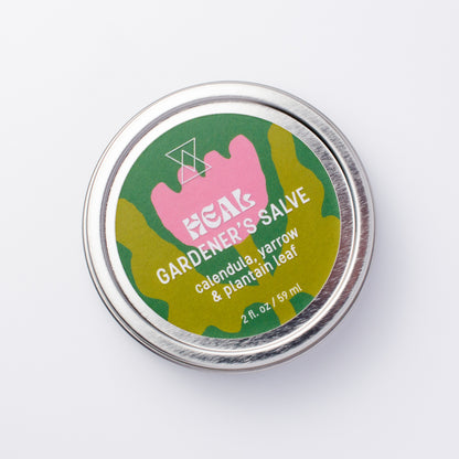 A round silver metal tin of Heal Gardener's Salve on a clean white background. The label features a pink flower shape over a green background, with white text reading 'HEAL GARDENER'S SALVE' and 'calendula, yarrow & plantain leaf'.