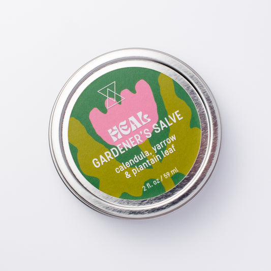 A round silver metal tin of Heal Gardener's Salve on a clean white background. The label features a pink flower shape over a green background, with white text reading 'HEAL GARDENER'S SALVE' and 'calendula, yarrow & plantain leaf'.