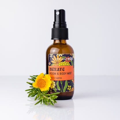 An amber glass spray bottle labeled 'HEKATE ROOM & BODY MIST' with a black pump, standing on a white surface against a light gray background, with a yellow flower and green sprigs placed in front.