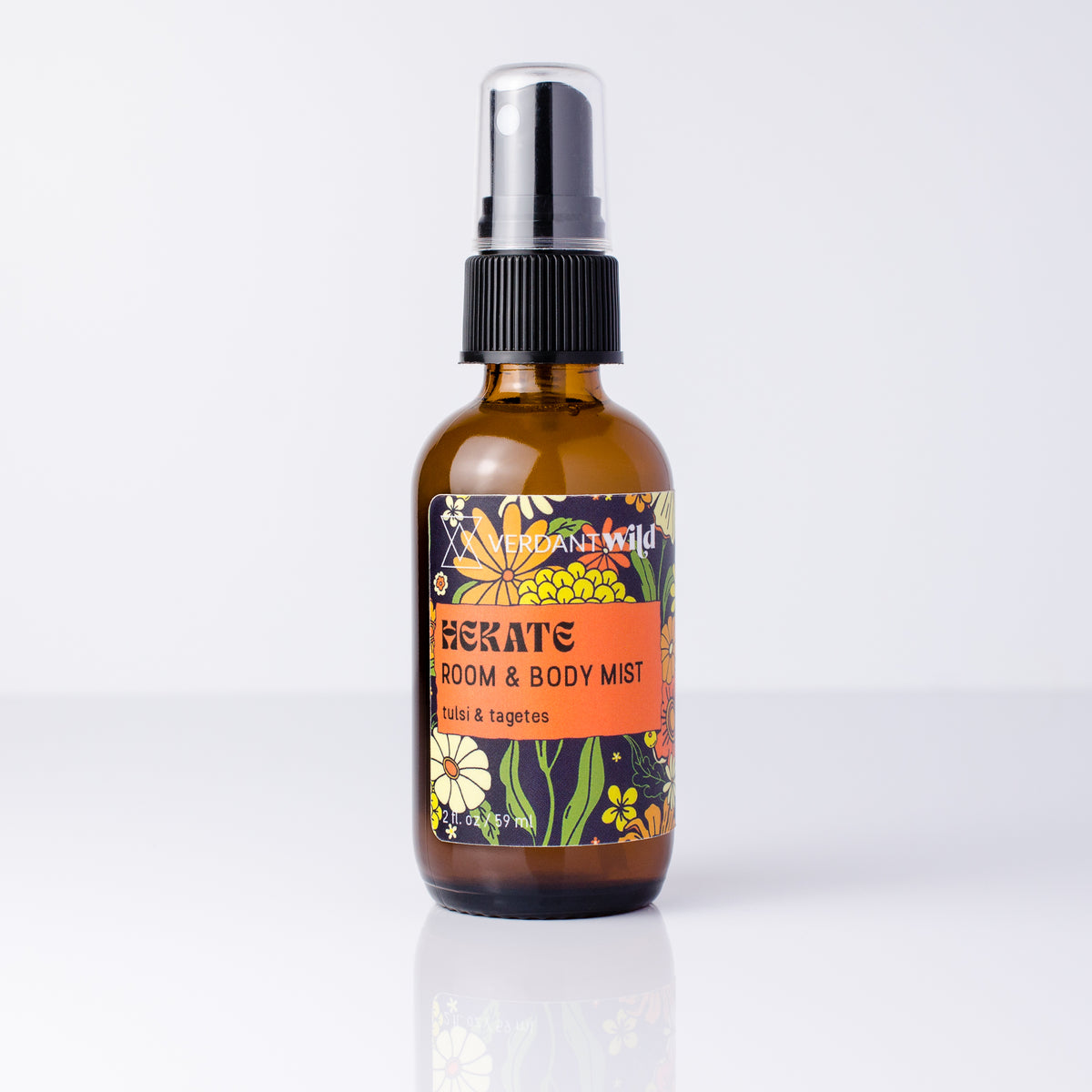 An amber glass room and body mist spray bottle with a black spray cap and a colorful floral label, shown on a clean white background with a soft reflection.