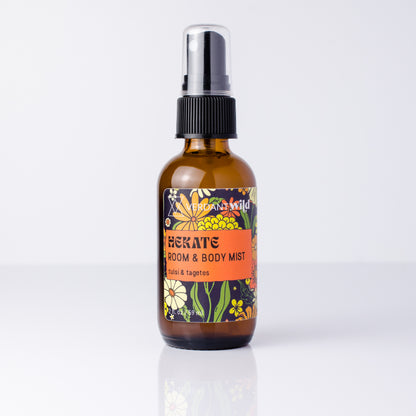 An amber glass room and body mist spray bottle with a black spray cap and a colorful floral label, shown on a clean white background with a soft reflection.