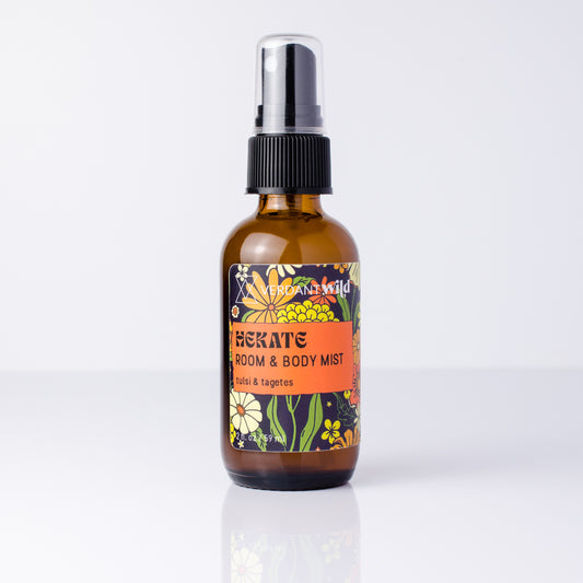 An amber glass room and body mist spray bottle with a black spray cap and a colorful floral label, shown on a clean white background with a soft reflection.