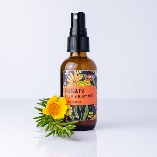 An amber glass spray bottle labeled 'HEKATE ROOM & BODY MIST' with a black pump, standing on a white surface against a light gray background, with a yellow flower and green sprigs placed in front.