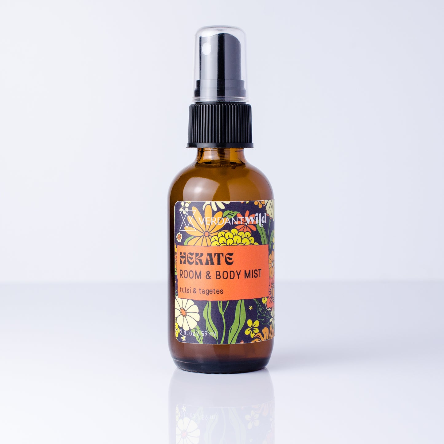 An amber glass spray bottle of Tulsi Room & Body Mist by Fragrance Wild, featuring a black spray cap and a colorful floral label, set against a clean white background with a soft reflection.