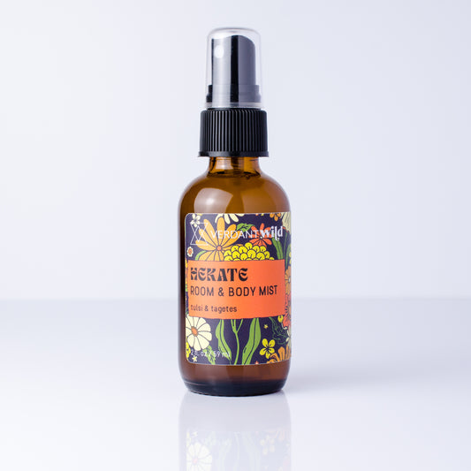 An amber glass spray bottle of Tulsi Room & Body Mist by Fragrance Wild, featuring a black spray cap and a colorful floral label, set against a clean white background with a soft reflection.