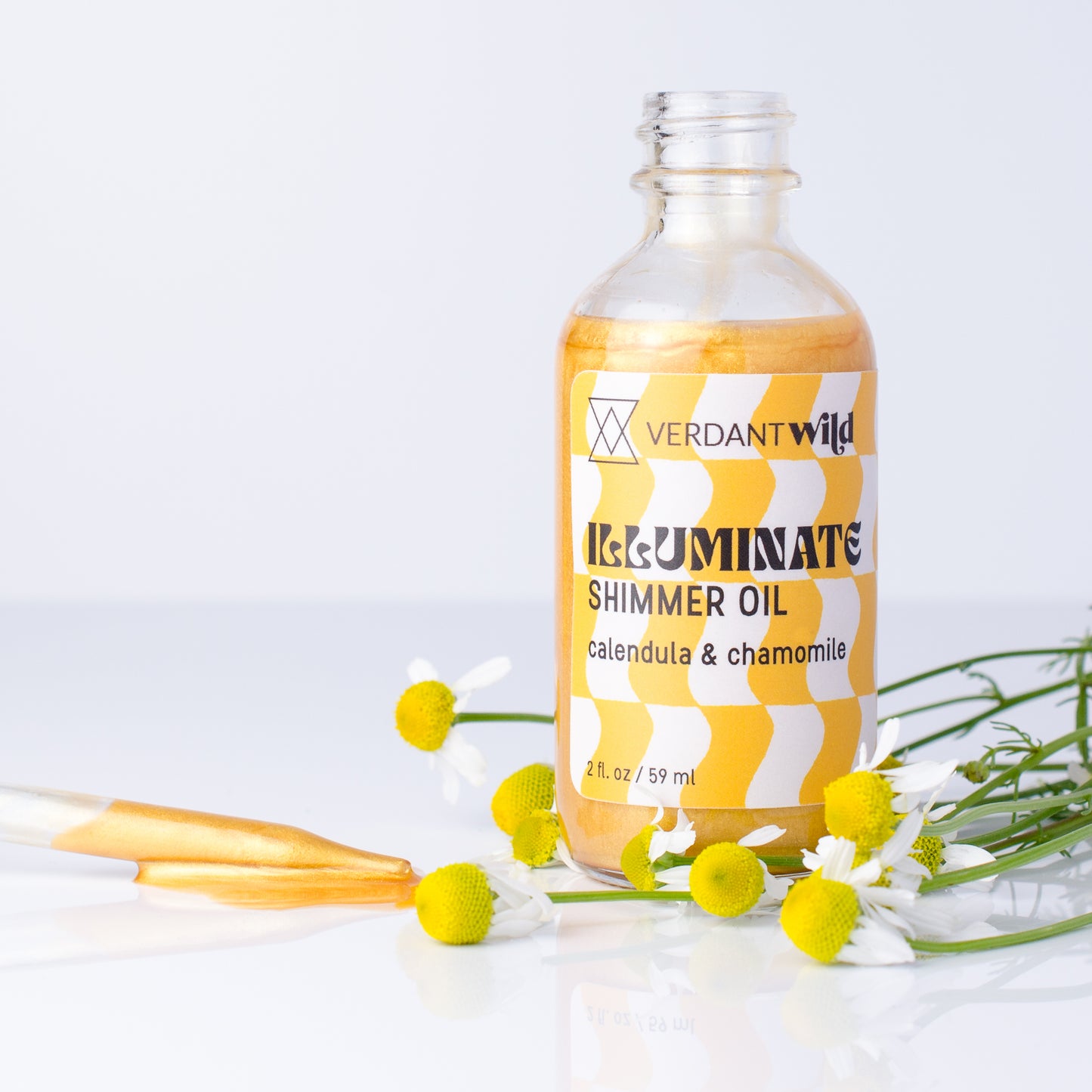 A clear glass bottle of Verdant Wild Illuminate Shimmer Oil with a yellow and white striped label, styled with chamomile flowers on a reflective white surface.