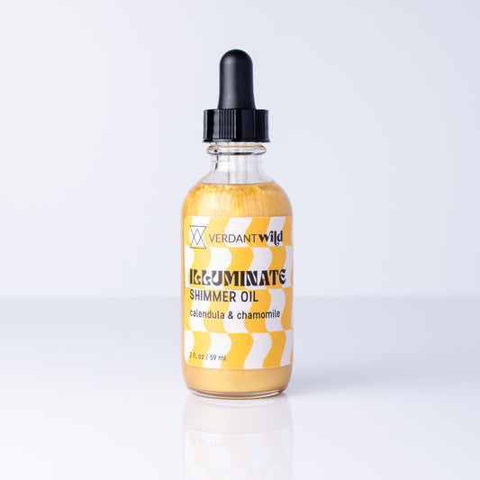 A bottle of Verdant Wild Illuminate Shimmer Oil with a black dropper cap, filled with shimmering golden-yellow oil, set against a clean white background with a soft reflection.