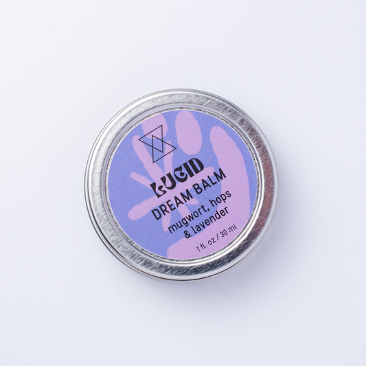 A round silver metal tin of LUCID Dream Balm on a clean white background. The label is lavender purple with black text reading 'mugwort, hops & lavender'.