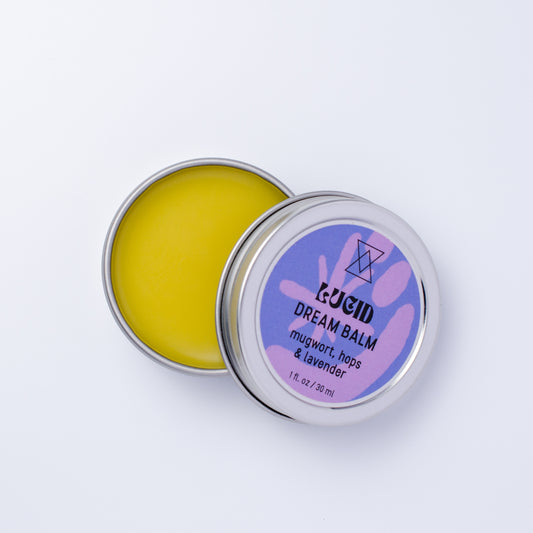An open round metal tin of Lucid Dream Balm showing the yellow balm inside, with the lid leaning against it. The lid label is purple with white text reading 'LUCID DREAM BALM mugwort, hops & lavender'.