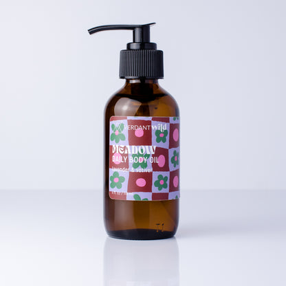 An amber glass pump bottle of Verdant Wild Meadow Daily Body Oil with a black pump and a colorful patterned label, set against a clean white background.