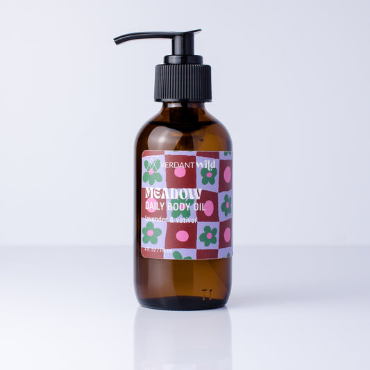 An amber glass pump bottle of Verdant Wild Meadow Daily Body Oil with a black pump and a colorful patterned label, set against a clean white background.
