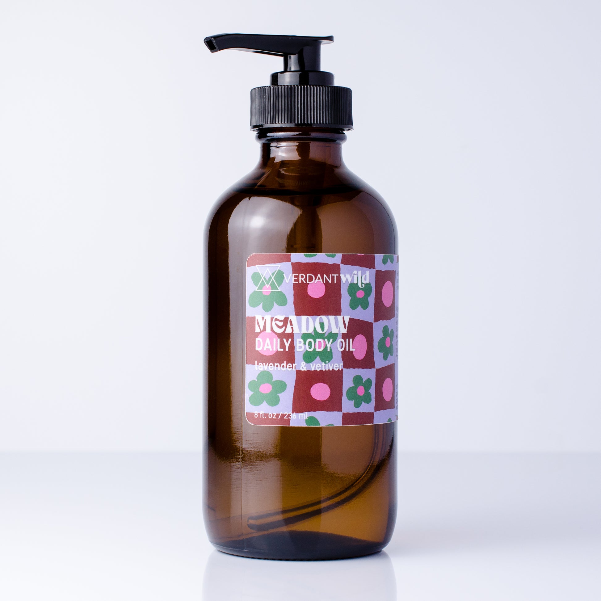 An amber glass pump bottle of Meadow Daily Body Oil by Verdant Wild, featuring a colorful floral patterned label and a black pump dispenser, set against a clean white background.