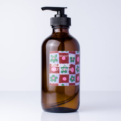 An amber glass pump bottle of Meadow Daily Body Oil by Verdant Wild, featuring a colorful floral patterned label and a black pump dispenser, set against a clean white background.
