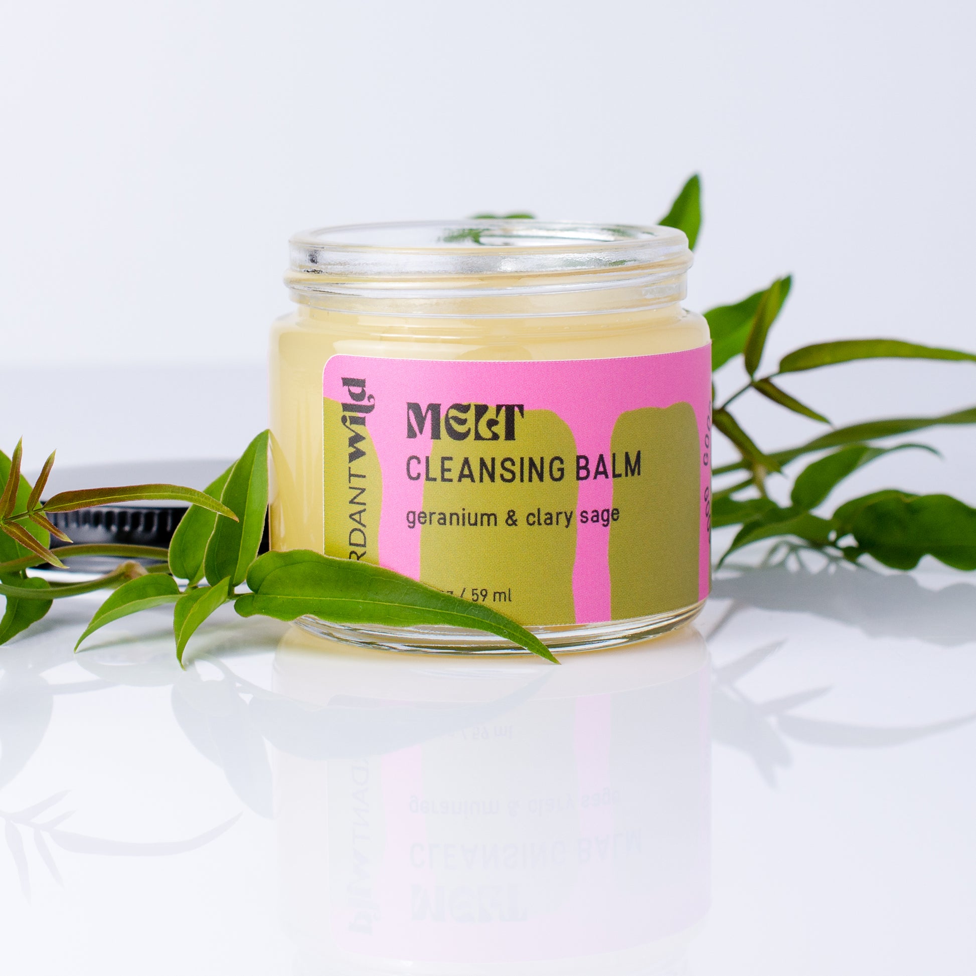 A glass jar of Melt Cleansing Balm by Brand Wild, filled with pale yellow balm and featuring a pink and olive-green label, styled with green leaves on a reflective white surface.