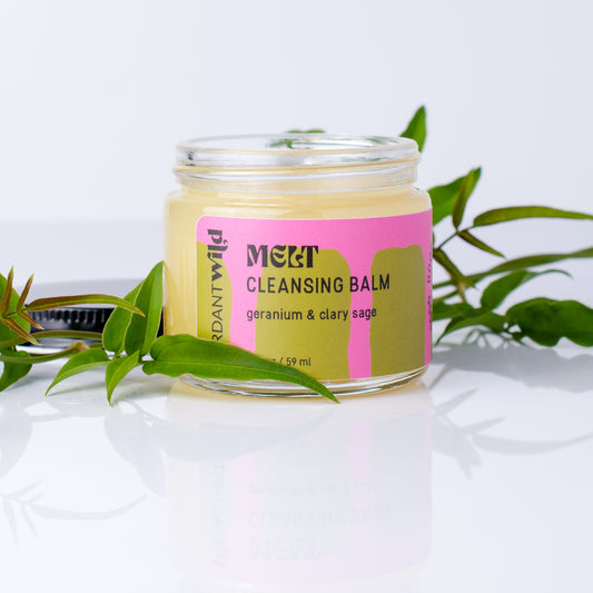 A glass jar of Melt Cleansing Balm by Brand Wild, filled with pale yellow balm and featuring a pink and olive-green label, styled with green leaves on a reflective white surface.