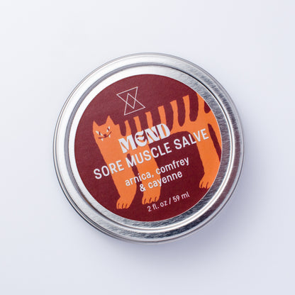 A round silver metal tin of MEND Sore Muscle Salve, featuring a dark red label with an orange cartoon cat and white text, set against a plain white background.