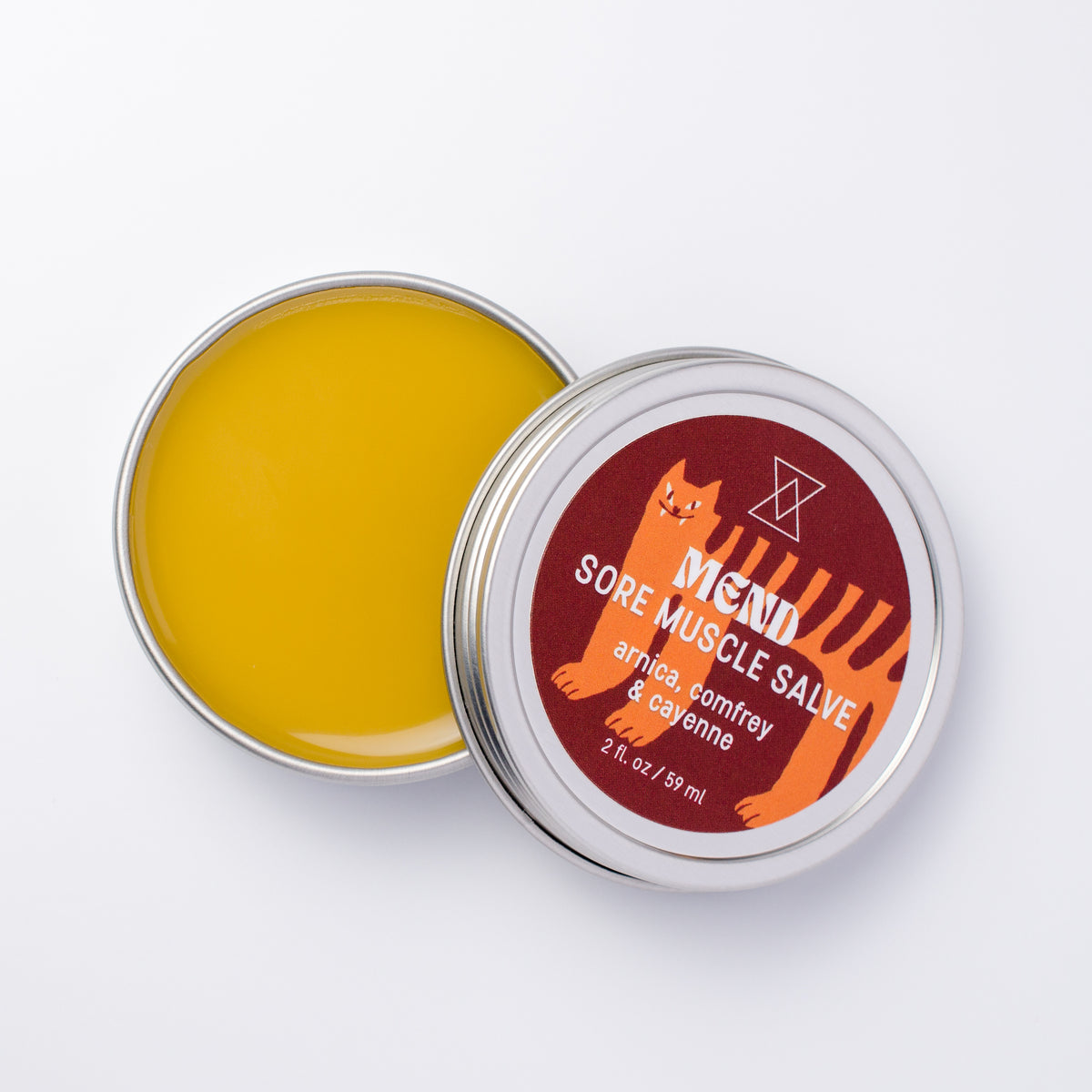 An open round metal tin of Mend Sore Muscle Salve showing the orange balm inside, with the lid placed beside it. The lid features a dark red label with an orange cat illustration and white text.