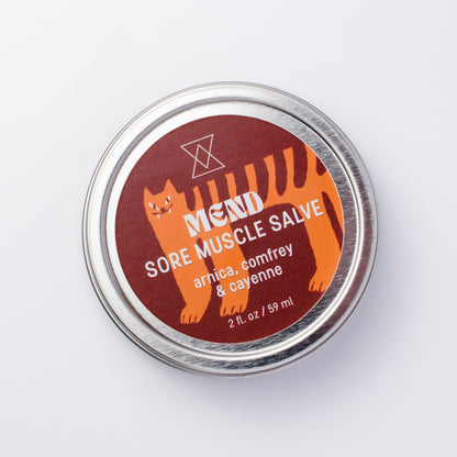 A round silver metal tin of MEND Sore Muscle Salve, featuring a dark red label with an orange cartoon cat and white text, set against a plain white background.