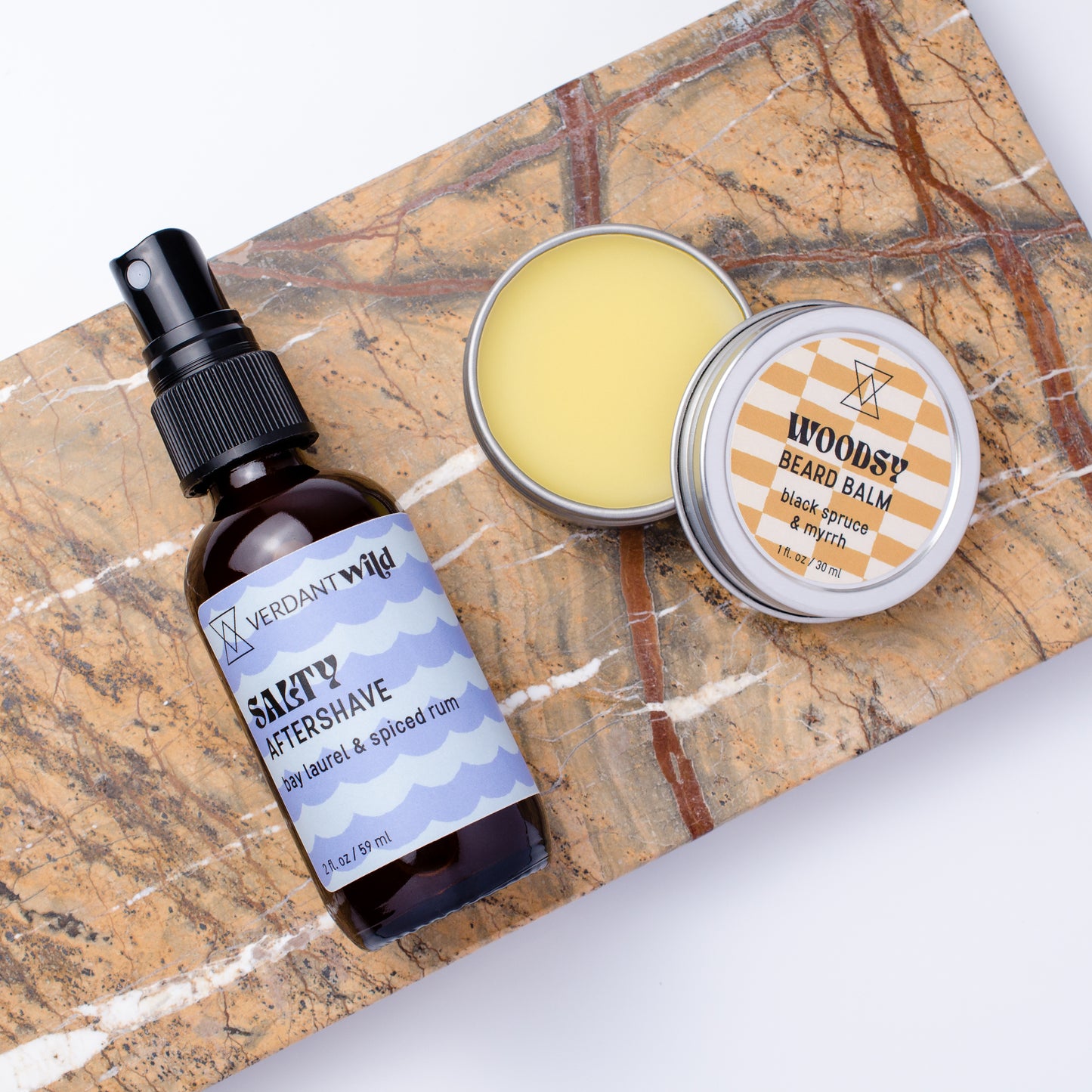 A grooming set featuring a dark amber spray bottle of Verdant Wild Salty Aftershave and a round tin of Woodsy Beard Balm, arranged on a brown and white marble slab against a white background.