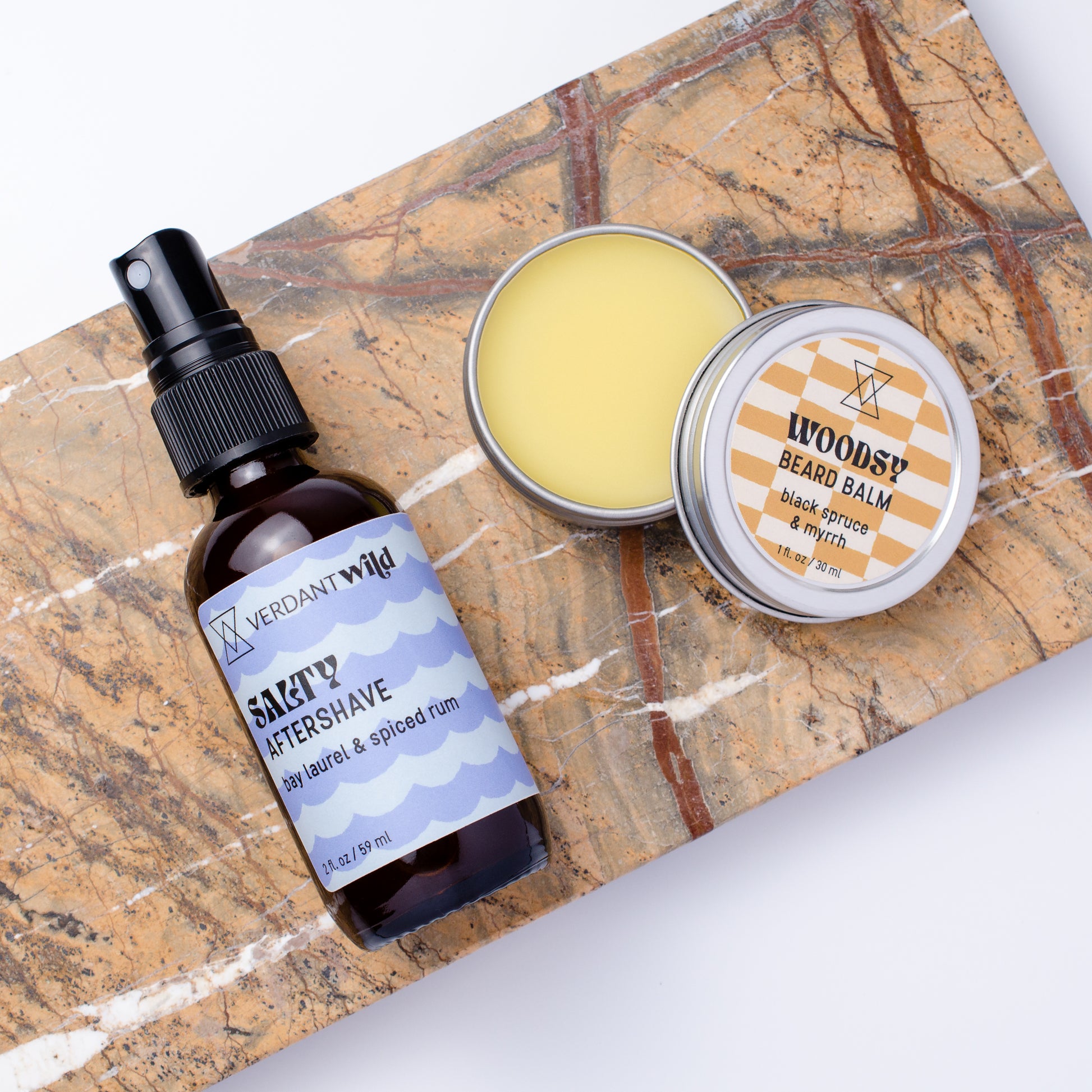 A grooming set featuring a dark amber spray bottle of Verdant Wild Salty Aftershave and a round tin of Woodsy Beard Balm, arranged on a brown and white marble slab against a white background.
