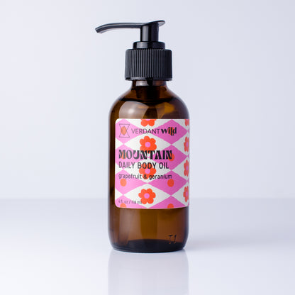 A 4 oz amber glass pump bottle of Verdant Wild Mountain Daily Body Oil with a pink and white patterned label, set against a clean white background.