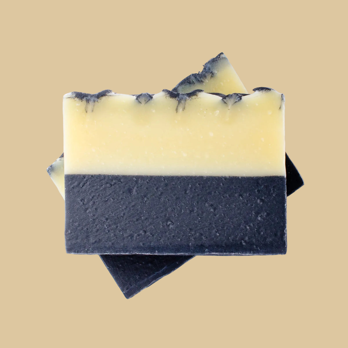 Two rectangular handmade soap bars stacked on top of each other, with one bar fully visible and the other partially hidden behind it. The front soap bar is split horizontally into two distinct sections: a pale creamy yellow top half and a dark charcoal gray bottom half.