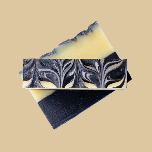 Two artisanal soap bars stacked diagonally on a solid beige background. The top bar features a marbled pattern of black and pale yellow.