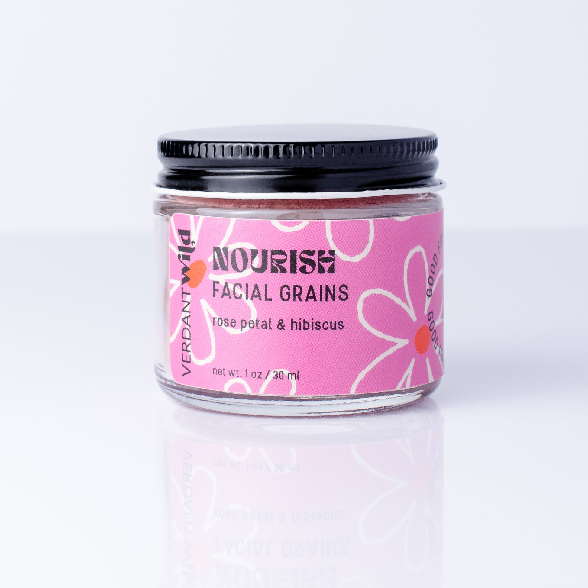 A jar of Verdant Wild Nourish Facial Grains in rose petal and hibiscus, featuring a pink label with white floral designs and a black metal lid, set against a clean white background with a soft reflection.