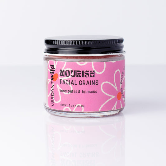 A small glass jar of Verdant Wild Nourish Facial Grains in rose petal & hibiscus, featuring a pink label with white floral patterns and a black screw-on lid, set against a clean white background with a soft reflection.