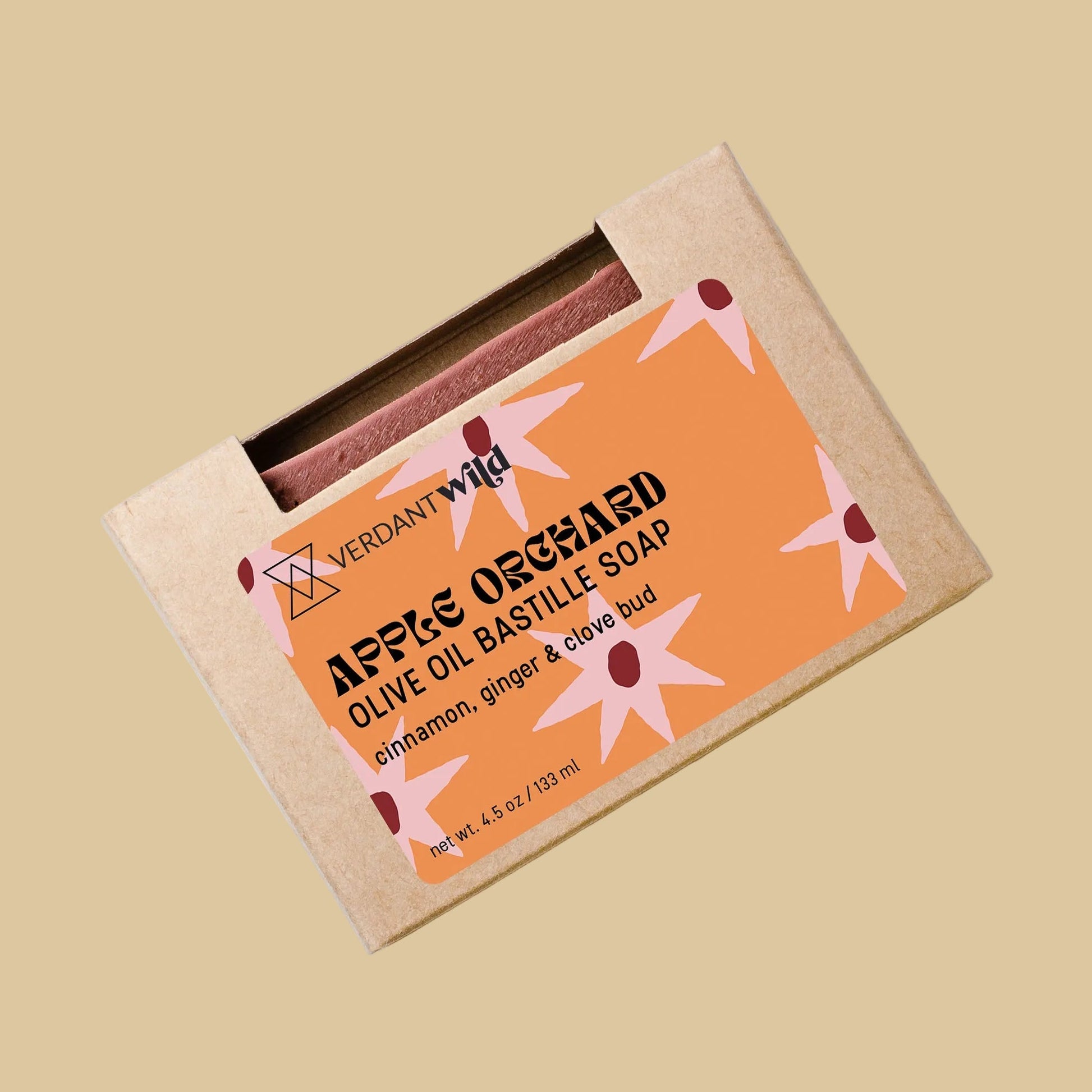 A bar of soap in a kraft paper sleeve with a peach-colored label that reads 'APPLES OLIVE OIL BASTILLE SOAP' and 'cinnamon, ginger & clove bud.' 
