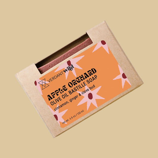 A bar of soap in a kraft paper sleeve with a peach-colored label that reads 'APPLES OLIVE OIL BASTILLE SOAP' and 'cinnamon, ginger & clove bud.' 