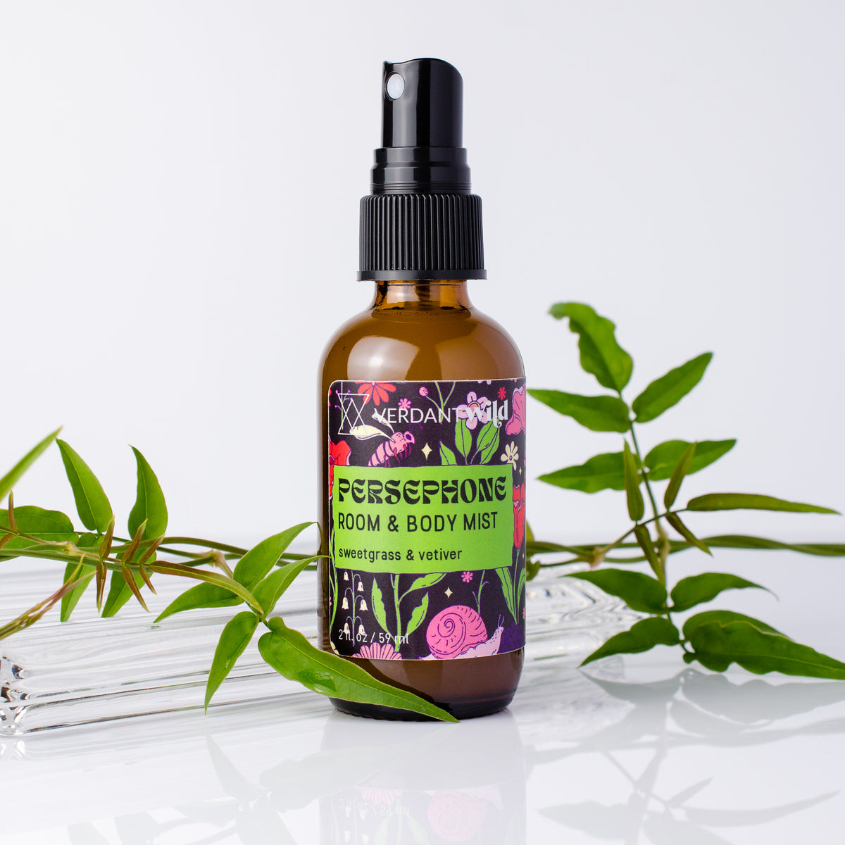 A bottle of Persephone Room & Body Mist with sweetgrass and vetiver scent, styled with green leaves on a reflective white surface against a white background.