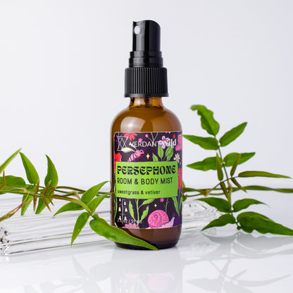 A bottle of Persephone Room & Body Mist with sweetgrass and vetiver scent, styled with green leaves on a reflective white surface against a white background.