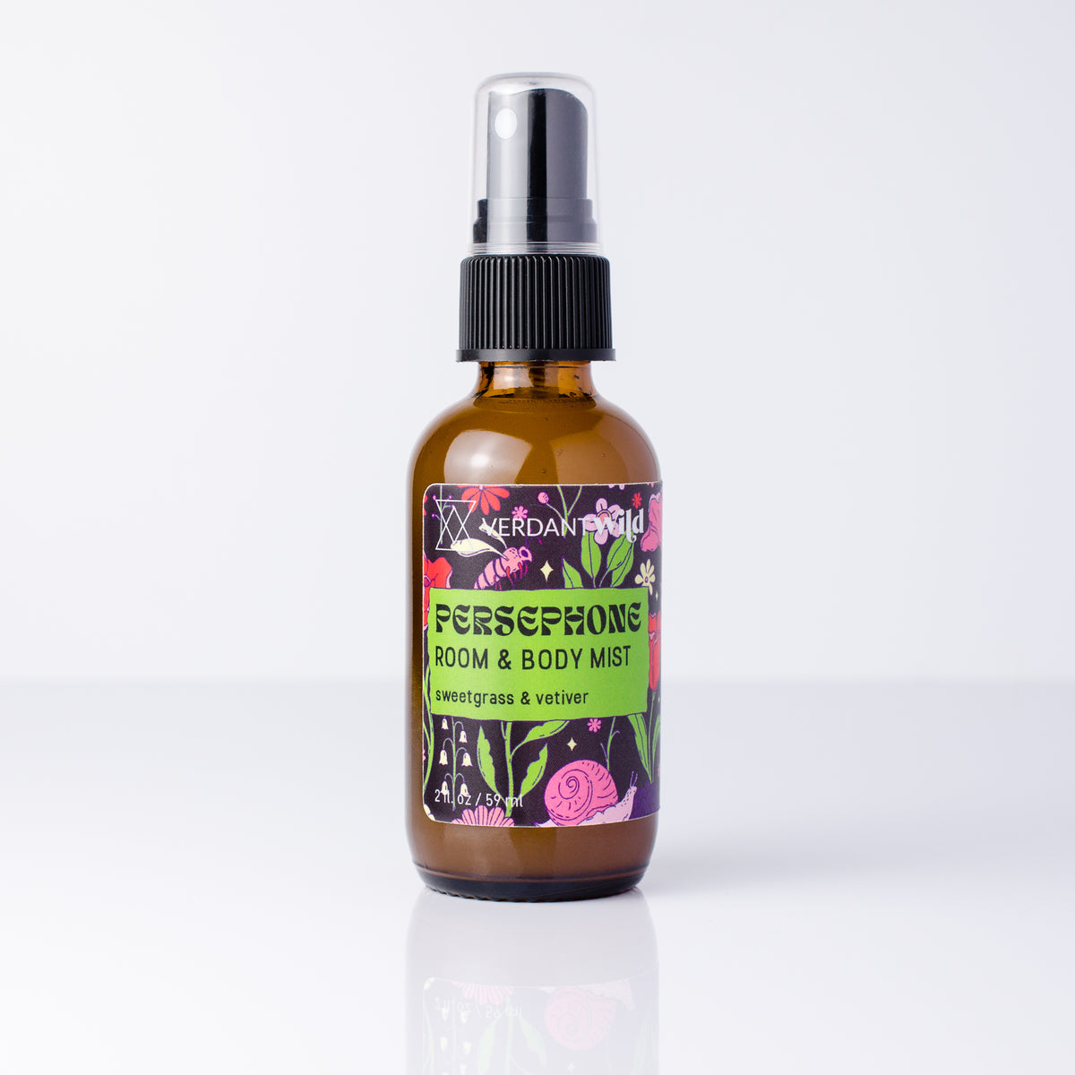 An amber glass room and body mist spray bottle with a black spray cap and a colorful floral label, standing on a white reflective surface.