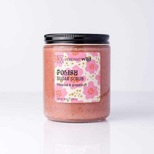 A jar of Verdant Wild Sugar Scrub in hibiscus & grapefruit scent, featuring pink sugar crystals inside a clear glass jar with a black lid and a floral pink label.