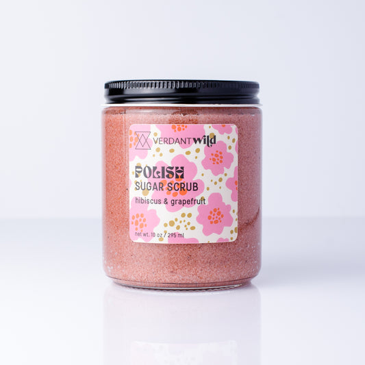 A jar of Verdant Wild Polish Sugar Scrub in hibiscus & grapefruit, featuring pink scrub inside a clear glass jar with a black lid and a pink floral label.