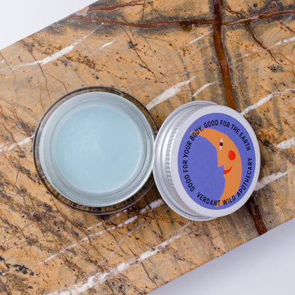 An open round glass jar containing a pale blue cream, placed on a brown marbled surface next to the silver lid. The lid has a colorful label with a crescent moon illustration and the text 'GOOD FOR YOUR BODY, GOOD FOR THE EARTH VERDANT WILD'.