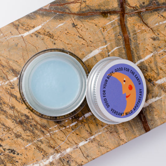 An open round jar of light blue face cream sits on a brown and beige marbled surface next to its lid, which features a colorful label with a crescent moon illustration and the text 'GOOD FOR YOUR BODY, GOOD FOR THE EARTH VERDANT WILD APOTHECARY'.