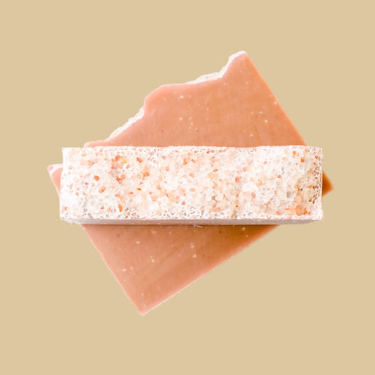 A rectangular bar of soap with a speckled, textured top layer resting diagonally on top of a smoother, solid-colored layer, set against a clean background.