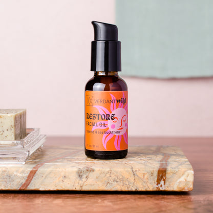 A small amber glass pump bottle labeled Verdant Wild Restore Facial Oil stands on a marble slab. A bar of soap sits on a glass dish to the left, and the background is softly blurred in warm neutral tones.