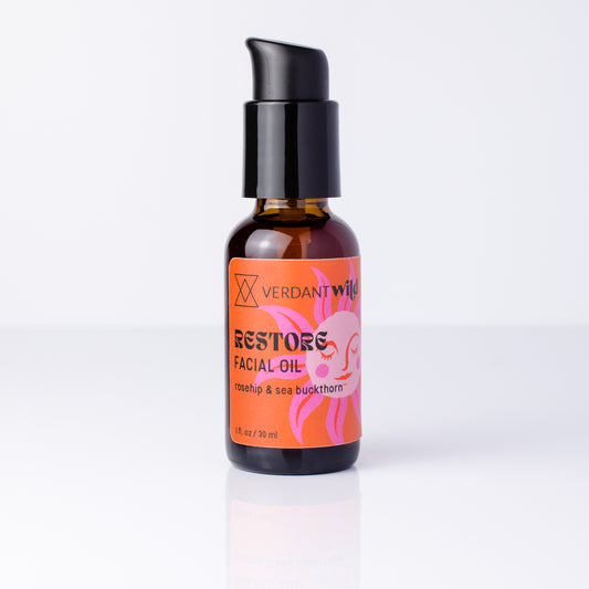 A small amber glass bottle of Verdant Wild Restore Facial Oil with a black pump dispenser and a bright orange label featuring pink and black text, set against a clean white background.