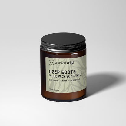 A jar candle with a dark amber glass body, a black metal lid, and a grey green label reading 'Verdant Wild DEEP ROOTS WOOD WICK SOY CANDLE'. The candle is photographed on a light gray studio background with a soft shadow to the right.