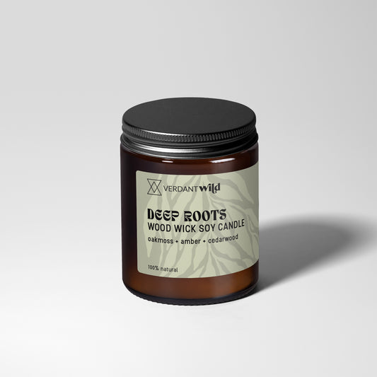 A jar candle with a dark amber glass body, a black metal lid, and a grey green label reading 'Verdant Wild DEEP ROOTS WOOD WICK SOY CANDLE'. The candle is photographed on a light gray studio background with a soft shadow to the right.