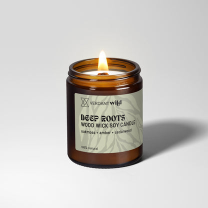 A lit amber glass jar candle with a sage green label reading Verdant Wild Deep Roots Wood Wick Soy Candle, set against a clean light gray background.