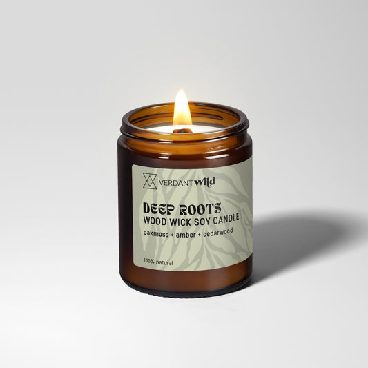 A lit amber glass jar candle with a sage green label reading Verdant Wild Deep Roots Wood Wick Soy Candle, set against a clean light gray background.